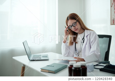 A woman in a white lab coat is sitting at a desk with a laptop 120428340