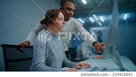 Female hotline operator in headset monitors data on computer with African American colleague Female hotline operator in headset monitors data on computer with African American colleague 120428891