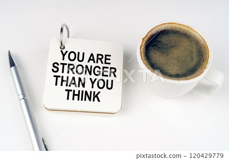 On a white surface, a cup of coffee, a pen and a notepad with the inscription - You Are Stronger Than You Think 120429779