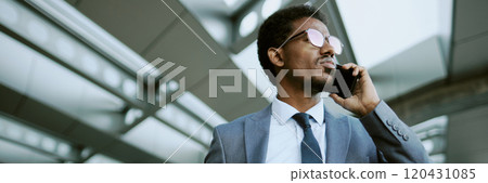 A professional businessman in a suit standing outside and talking on smartphone, reflecting light from his glasses while looking ahead, steel architectural details in background 120431085
