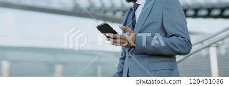 Businessman holding smartphone and standing in modern glass building looking confident and professional while focusing on device without looking straight at camera 120431086