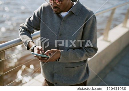 Man standing by waterfront rail while engrossed in browsing smartphone. Capturing casual moment at urban outdoor location 120431088