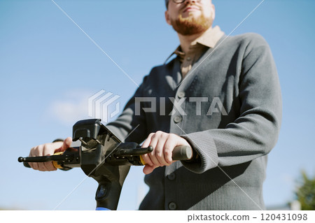 Young man riding electric scooter outside during sunny day wearing a grey cardigan against clear blue sky. Captured in urban setting with relaxed and casual vibe 120431098