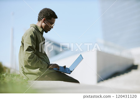 Man wearing casual attire sitting on grassy area using laptop for remote work outdoors in sunny environment showing modern work flexibility Man wearing casual attire sitting on grassy area using laptop for remote work outdoors in sunny environment showing modern work flexibility 120431120