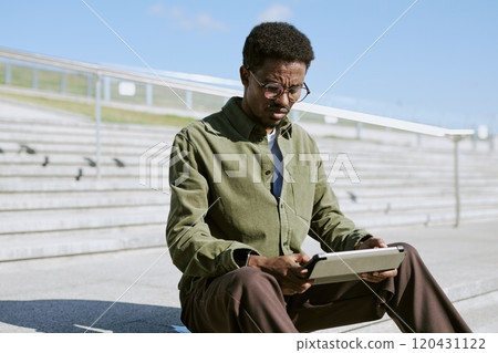 African American man sitting outdoors while using a tablet. Steps and railing are in the background, providing a casual atmosphere African American man sitting outdoors while using a tablet. Steps and railing are in the background, providing a casual atmosphere 120431122