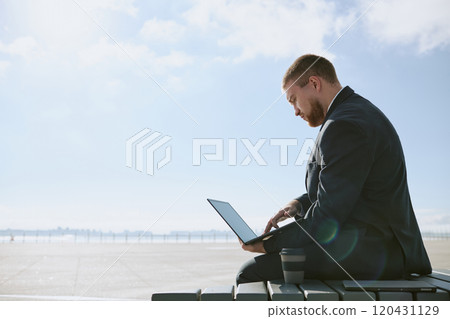 Businessman dressed in suit working on laptop while sitting at airport terminal. Clear sky in background, with coffee cup placed beside him Businessman dressed in suit working on laptop while sitting at airport terminal. Clear sky in background, with coffee cup placed beside him 120431129