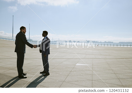 Two business professionals shaking hands standing in a spacious outdoor area surrounded by large cityscape on horizon. Background features a vast, open space under clear blue sky Two business professionals shaking hands standing in a spacious outdoor area surrounded by large cityscape on horizon. Background features a vast, open space under clear blue sky 120431132