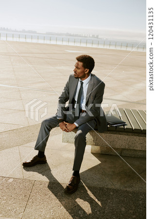 Businessperson in gray suit sitting on bench with ocean background Staring into distance and holding hands together Man with neatly groomed facial hair 120431135