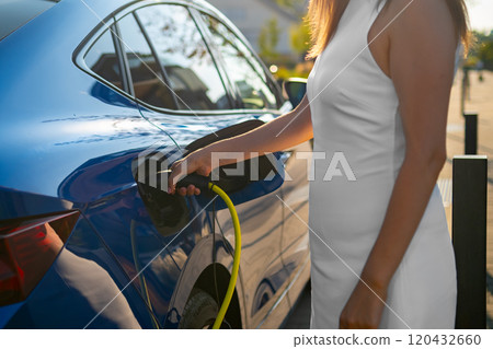 Close up woman in a white dress connecting an electric car to a charging station on a city street Close up woman in a white dress connecting an electric car to a charging station on a city street 120432660