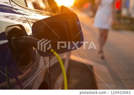Close up electric car charging, while a woman in a white dress is walking beside it at sunset.  120432661