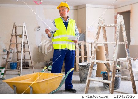 Portrait of worker in a yellow vest with documents in his hands in renovated cottage Portrait of worker in a yellow vest with documents in his hands in renovated cottage 120438520