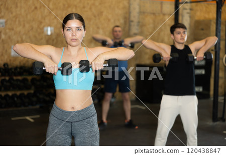 Motivated young woman practicing exercises with dumbbells near other people in gym 120438847