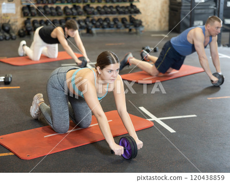 Motivated young woman practicing exercises with abs roller near other people in gym 120438859