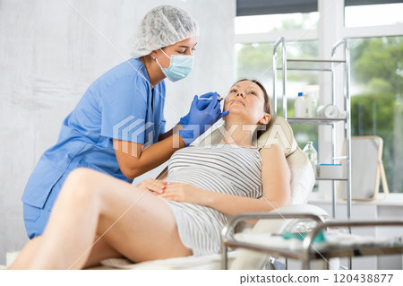 Doctor in disposable mask performs procedure of lip augmentation of girl patient in clinic. Doctor in disposable mask performs procedure of lip augmentation of girl patient in clinic. 120438877