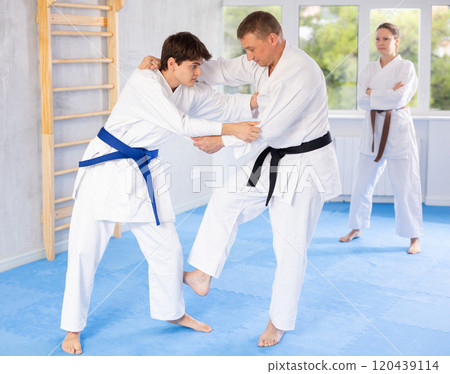 Judo or jiu-jitsu classes - two men practicing grabbing and throwing on sports mats under the guidance of trainer 120439114