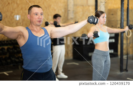 Motivated man practicing exercises with dumbbells near other people in gym Motivated man practicing exercises with dumbbells near other people in gym 120439170