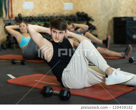 Motivated sporty young man lying on his back and training core muscles during group training class in CrossFit center 120439182
