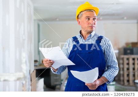 Portrait of worker in blue overall with documents in his hands in renovated cottage 120439273