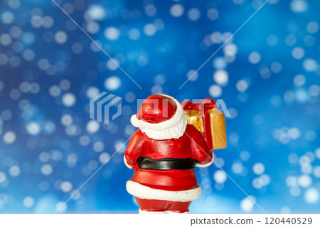 Cheerful Santa Claus figurine holding a golden gift box, set against a vibrant blue background with snowy bokeh effects 120440529