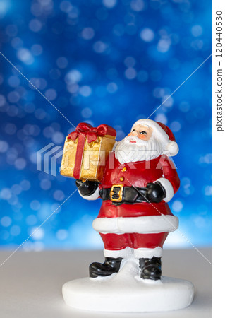 Cheerful Santa Claus figurine holding a golden gift box, set against a vibrant blue background with snowy bokeh effects 120440530