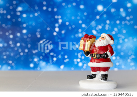 Cheerful Santa Claus figurine holding a golden gift box, set against a vibrant blue background with snowy bokeh effects 120440533
