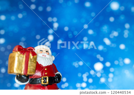 Cheerful Santa Claus figurine holding a golden gift box, set against a vibrant blue background with snowy bokeh effects 120440534