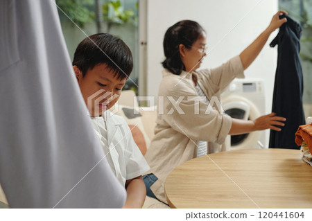 Boy Sitting While Woman Hanging Clothes Indoors Boy Sitting While Woman Hanging Clothes Indoors 120441604