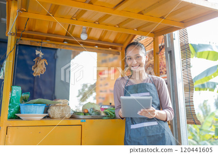 A Smiling Woman at a Food Stall Holding a Tablet, Showcasing Local Fresh Ingredients 120441605