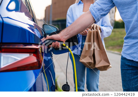 Person charges an electric car while another holds shopping bags at an urban parking lot Person charges an electric car while another holds shopping bags at an urban parking lot 120442419