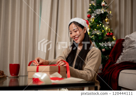 A smiling woman prepares Christmas presents in the living room, tying a red ribbon around a gift box A smiling woman prepares Christmas presents in the living room, tying a red ribbon around a gift box 120444111
