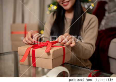 A close-up of a happy woman tying a red ribbon around a Christmas gift box in the living room. 120444116