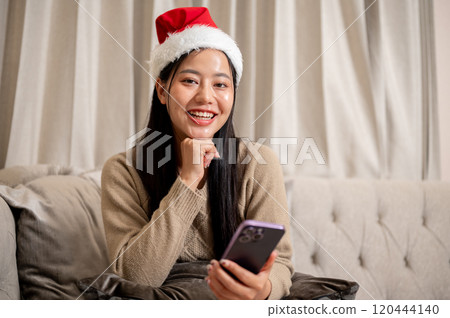 A beautiful Asian woman in cozy clothes and a Christmas hat sits on the sofa in the living room. A beautiful Asian woman in cozy clothes and a Christmas hat sits on the sofa in the living room. 120444140