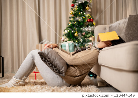 A sleepy woman rests a book on her face, falling asleep while reading, celebrating Christmas night. 120444149