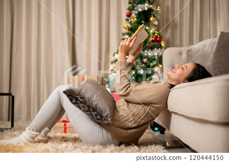 A side view of a happy woman enjoys reading a book in the living room during the Christmas night. 120444150