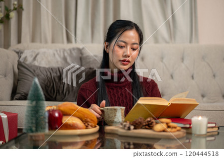 A beautiful Asian woman sits in her living room, reading a book during the Christmas holiday. 120444518