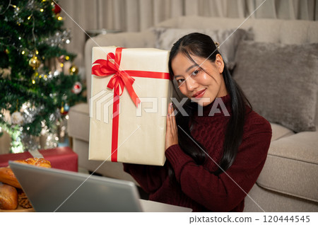 Beautiful Asian woman sits in her living room, smiling and showing the Christmas gift to the camera. Beautiful Asian woman sits in her living room, smiling and showing the Christmas gift to the camera. 120444545