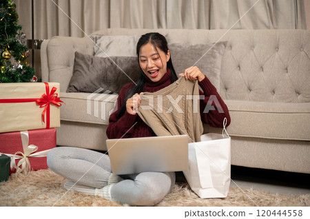 An excited Asian woman showing her Christmas present to the camera while having a video call. 120444558