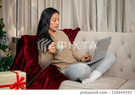An attractive Asian woman in warm cozy clothes using her laptop on the sofa in the Christmas night. An attractive Asian woman in warm cozy clothes using her laptop on the sofa in the Christmas night. 120444593