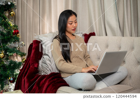 An attractive Asian woman in warm cozy clothes using her laptop on the sofa in the Christmas night. An attractive Asian woman in warm cozy clothes using her laptop on the sofa in the Christmas night. 120444594