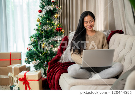 An attractive Asian woman in warm cozy clothes using her laptop on the sofa in the Christmas day. An attractive Asian woman in warm cozy clothes using her laptop on the sofa in the Christmas day. 120444595