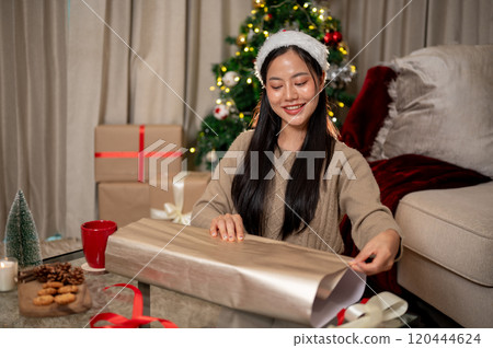 A charming, happy Asian woman in cozy clothes wraps a Christmas present in the living room. A charming, happy Asian woman in cozy clothes wraps a Christmas present in the living room. 120444624