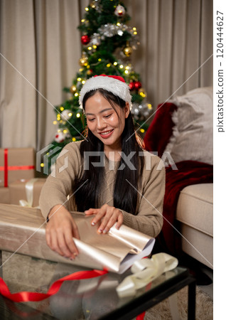 A charming, happy Asian woman in cozy clothes wraps a Christmas present in the living room. A charming, happy Asian woman in cozy clothes wraps a Christmas present in the living room. 120444627