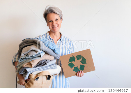 Gray haired woman with pile of clothes and recycling reusing signboard isolated on white background Gray haired woman with pile of clothes and recycling reusing signboard isolated on white background 120444917