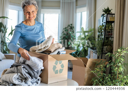Gray haired mature smiling woman pack clothes for recycling to cardboard box at home Gray haired mature smiling woman pack clothes for recycling to cardboard box at home 120444936