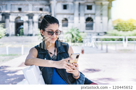Urban style girl sitting on a bench with her cell phone, An attractive girl sitting on the cell phone smiling, Girl sitting on a bench texting on her cell phone. Urban style girl sitting on a bench with her cell phone, An attractive girl sitting on the cell phone smiling, Girl sitting on a bench texting on her cell phone. 120445652