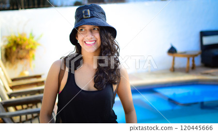 Pretty girl with hat smiling near a swimming pool, latin girl smiling by a swimming pool with muskoka chairs Pretty girl with hat smiling near a swimming pool, latin girl smiling by a swimming pool with muskoka chairs 120445666