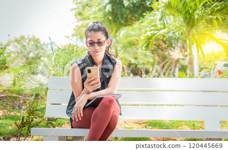 Girl sitting on a bench checking her cell phone, Happy woman sitting in a park texting on her cell phone, Woman sitting on a bench sending a text message Girl sitting on a bench checking her cell phone, Happy woman sitting in a park texting on her cell phone, Woman sitting on a bench sending a text message 120445669