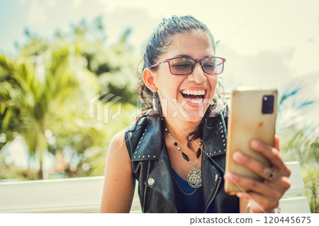 Happy girl with cell phone in a park. Young woman laughing at cell phone sitting on a bench. Happy girl in glasses with cell phone on a bench in the street Happy girl with cell phone in a park. Young woman laughing at cell phone sitting on a bench. Happy girl in glasses with cell phone on a bench in the street 120445675