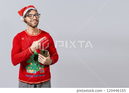 Christmas man holding phone pointing to the side. Smiling young man in christmas hat with cellphone pointing at promo. Christmas man concept with cell phone pointing to the side 120446185