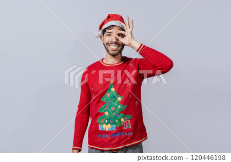 Funny young man in christmas hat looking through fingers. Funny christmas guy making gesture and looking through fingers, Man in christmas hat making gesture looking through fingers 120446198